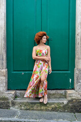 Fototapeta na wymiar Beautiful redhead woman in long colorful dress standing against a large green colored door.