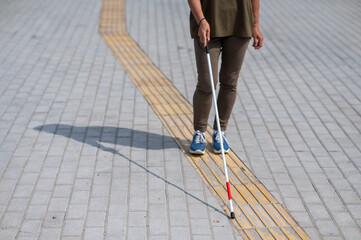 Close-up of the legs of an elderly blind woman with a cane at a tactile tile. - obrazy, fototapety, plakaty