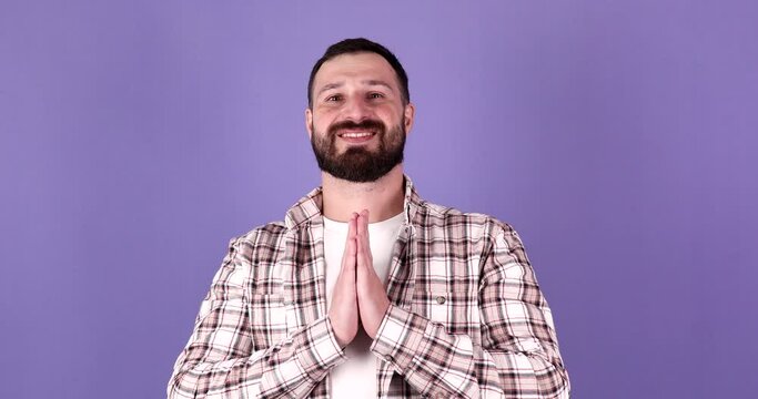 Portrait bearded man thank gesture with connected hands and reverence smiling posing isolated on purple background. Calmness happy male arms namaste praying yoga position with positive emotion.