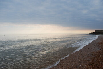 Incoming Tide Atlantic Ocean Sea Ebbing and Flowing on Smooth Natural Stone Pebble Beach in England with Misty Stormy Horizon with Setting Sun Casting Pinks Oranges Peeking Through Stormfront 