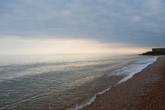 Incoming Tide Atlantic Ocean Sea Ebbing and Flowing on Smooth Natural Stone Pebble Beach in England with Misty Stormy Horizon with Setting Sun Casting Pinks Oranges Peeking Through Stormfront 