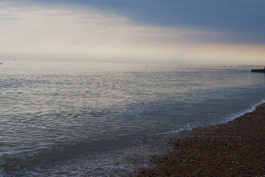 Receding Tide Atlantic Ocean Sea Ebbing and Flowing on Smooth Natural Stone Rock Pebble Beach Coastline Shore in England UK Britain with Misty Stormy Horizon with Setting Sun Casting Pinks Oranges 