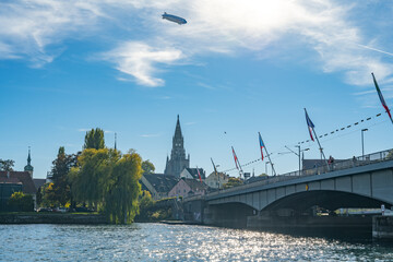 Zeppelin flying over the city on a sunny day with tourists walking across a bridge at the Steigenberger Peninsula in the foreground. (Konstanz), Lake Constance (Bodensee), Baden-Württemberg, Germany. © Michael