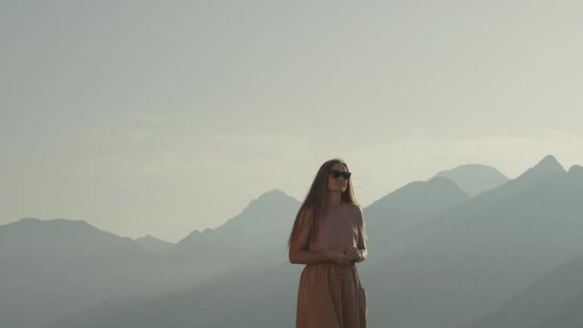 Amidst the mountains in the soft cinematic light of the setting sun, a young woman looks around, she is captivated by the views. The concept of a new life in travel.