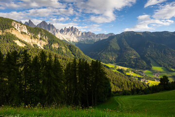 Picture of colorful nature, forest and meadows, against background of sky and mountains, in Val de Funes region, Dolomites. In distance are lonely rural houses of local residents