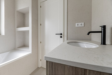 Image of the bathroom with cream-colored tiles, matching countertop, furniture with wooden drawers, black taps, white tub with built-in niche