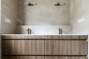 Front image of bathroom with cream marble tiles, matching countertop, wooden cabinet with drawers and gold taps with wall lights