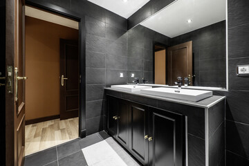 Tiled bathroom with black slate tiles, individual white porcelain sinks on black cabinet with mirror built into the wall