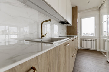 Modern open kitchen with white metal and glass retractable screen, gold metal taps and furniture handles, shiny stone imitation countertop and light wood furniture to match the wooden floor