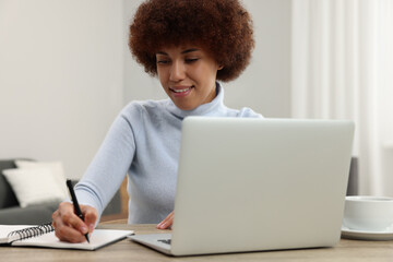 Beautiful young woman using laptop and writing in notebook at wooden desk in room