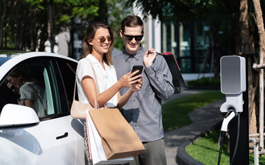 Young couple travel with EV electric car charging in green sustainable city outdoor garden in...
