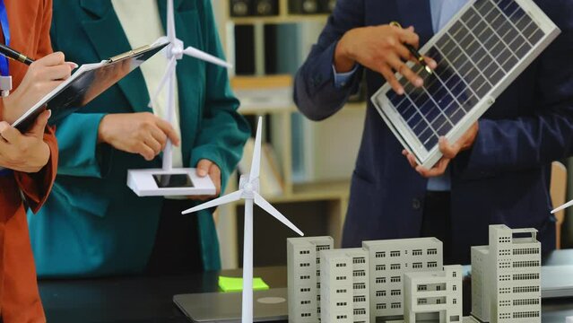 Four asian businesspeople professionals discuss renewable energy in office, showing solar panel and wind turbines with tower building model, apartment. modern business with environmental, clean energy