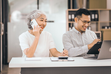 An older mature woman shows something to asian man on a laptop. They smile and work together in an office. They look happy and focused.