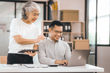 An older mature woman shows something to asian man on a laptop. They smile and work together in an office. They look happy and focused.