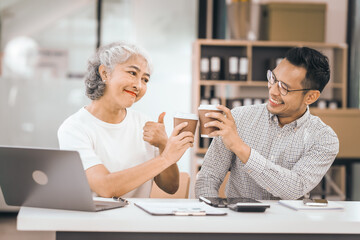 An older mature woman shows something to asian man on a laptop. They smile and work together in an office. They look happy and focused.