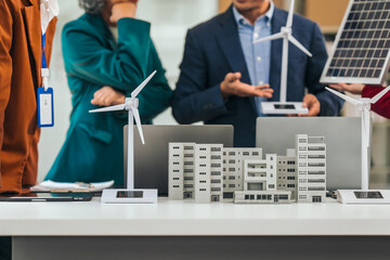 Four asian businesspeople professionals discuss renewable energy in office, showing solar panel and wind turbines with tower building model, apartment. modern business with environmental, clean energy