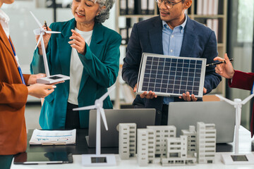 Four asian businesspeople professionals discuss renewable energy in office, showing solar panel and wind turbines with tower building model, apartment. modern business with environmental, clean energy - obrazy, fototapety, plakaty
