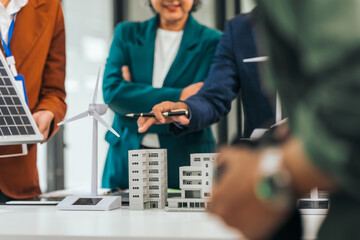 Four asian businesspeople professionals discuss renewable energy in office, showing solar panel and wind turbines with tower building model, apartment. modern business with environmental, clean energy