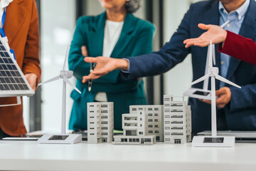 Four asian businesspeople professionals discuss renewable energy in office, showing solar panel and wind turbines with tower building model, apartment. modern business with environmental, clean energy