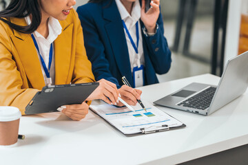 Two young asian business women work in an office. One uses a tablet and looks serious. The other smiles and types on a laptop.