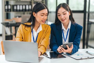 Fototapeta premium Two young asian business women work in an office. One uses a tablet and looks serious. The other smiles and types on a laptop.