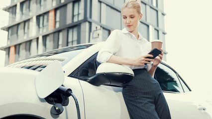 Progressive businesswoman leaning on electric car and charging station before driving around city...