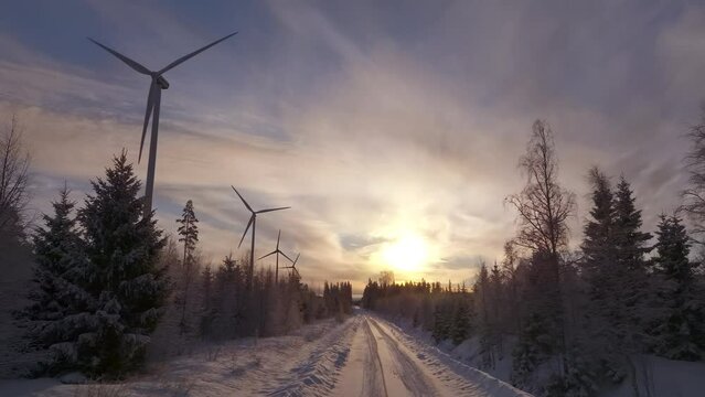 Scenic aerial video over forest winter road with windmills standing in row at the left side in forest. Large wind turbines with blades, drone camera fly forward.