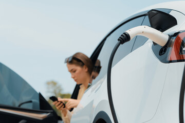 Young woman use smartphone to pay for electricity at public EV car charging station green city...