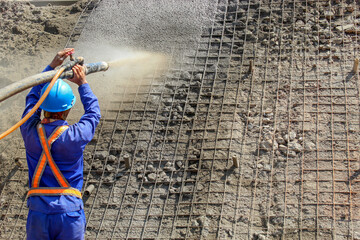 A worker is doing the shotcrete process, where the process is by firing concrete or spraying concrete into the ground covered by a wire mesh. This process is commonly used for retaining walls.