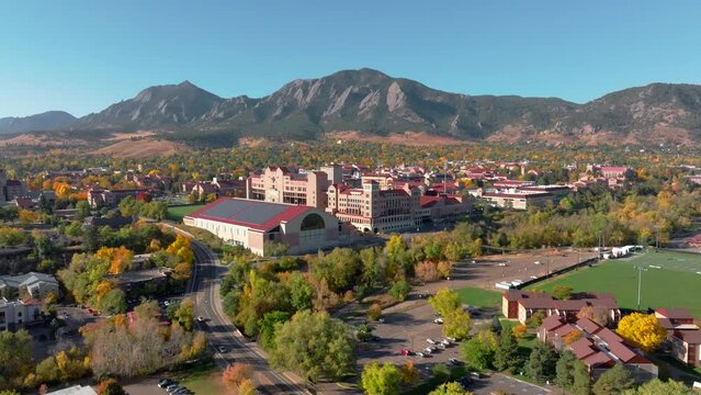 Aerial moving forward view of CU Boulder campus and stadium surrounded by green and yellow fall trees with the rocky flatiron mountains in the background in the front range of Colorado