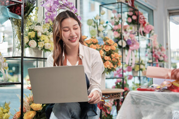 One young Asian female florist and SME entrepreneur in apron, works with laptop, online selling floral arrange in colorful flower shop store with beautiful bunch of blossoms, and e-commerce business.