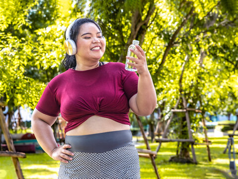 Fat woman wearing sportswear working out and wearing earphones listening to music relaxed breathing fresh air in a green forest  outdoors at the park in the morning with a fresh feeling.