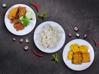 Ayam Penyet, fried tempe and tofu served with a plate of rice. A dish consists of fried chicken, tempeh, and tofu, served with spicy sambal, a slice of cucumber, cabbage, and lemon basil.
