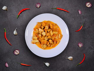 Spicy tempeh and tofu stew in coconut milk with garlics, shallots, and red chilis scattered around the plate. Tempe and tofu curry on a white plate with dark background. Indonesian homemade cuisine.
