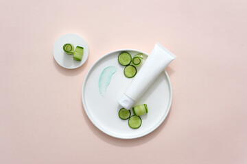 View from above, on a pink background, an unlabeled cosmetic tube, cucumber slices and two white plates. Homemade cosmetics with cucumber extract. Blank space for advertising.