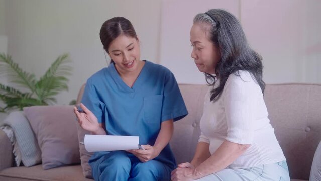 Asian caregiver nurse attentively inquires and records information from an elderly Asian woman, seated together on a living room sofa. She is dedicated to providing care and support to the elderly.