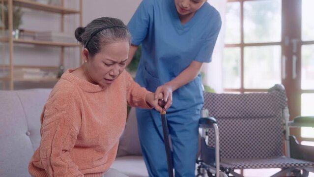 A compassionate young Asian female nurse helps an elderly Asian lady rise from the sofa using a cane in the living room of her home. The nurse offers sturdy support, promoting mobility.