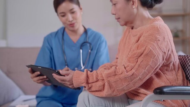 young Asian female nurse, sitting on the sofa, holds a tablet and provides a detailed explanation to an elderly Asian lady in a wheelchair in the living room.