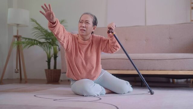 An elderly Asian woman, holding a walking cane, attempts to rise from the sofa but falls back, possibly due to a heart-related issue, in the living room. Her need for assistance is evident.