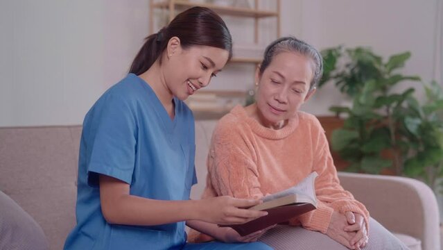 An Asian female caregiver reads a novel to an elderly Asian woman seated on a living room sofa. This image reflects the dedicated care and companionship offered to seniors at home.