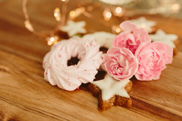 Gingerbread cookies in the shape of stars, merengue cookies and pink roses on a wooden table against a background of bokeh of New Year’s lights. Christmas background. Winter card. Delicious pastries
