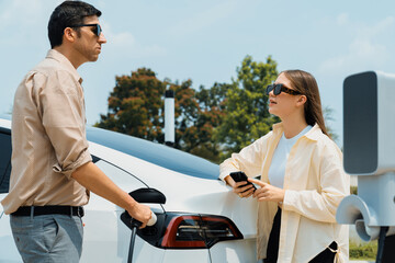 Young couple use smartphone to pay for electricity at public EV car charging station green city...