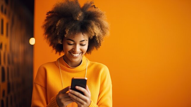 Happy beautiful young woman looking at her mobile phone and smiling. Pretty girl in casua standing on orange background, holding cell phone and reading funny message she received from boyfriend