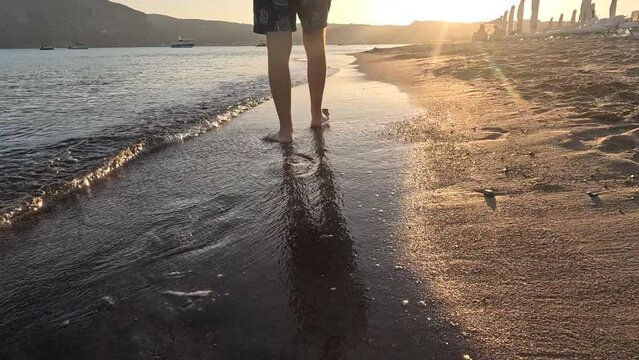 Man walking barefoot on sand, rear view. Male legs walking on a sandy shore with small flowing waves at sunset. Summer time island background in 4k resolution. 