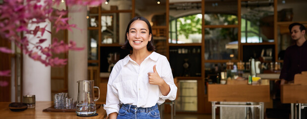 Confident asian businesswoman, showing thumbs up, standing near entrance of her cafe or restaurant, recommending place - obrazy, fototapety, plakaty