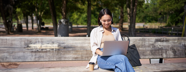 Portrait of korean woman sitting with laptop in park on bench, working outdoors, student doing homework, drinking takeaway coffee