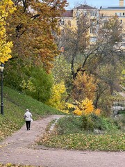 City park in ravine. Woman walks among autumn trees and leaf fall. Yellow house in the background. Road of sand leads down to the bridge. Golden autumn, beauty and tranquility of northern nature.