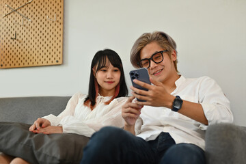 Happy young couple sitting on sofa and watching social media videos on smartphone