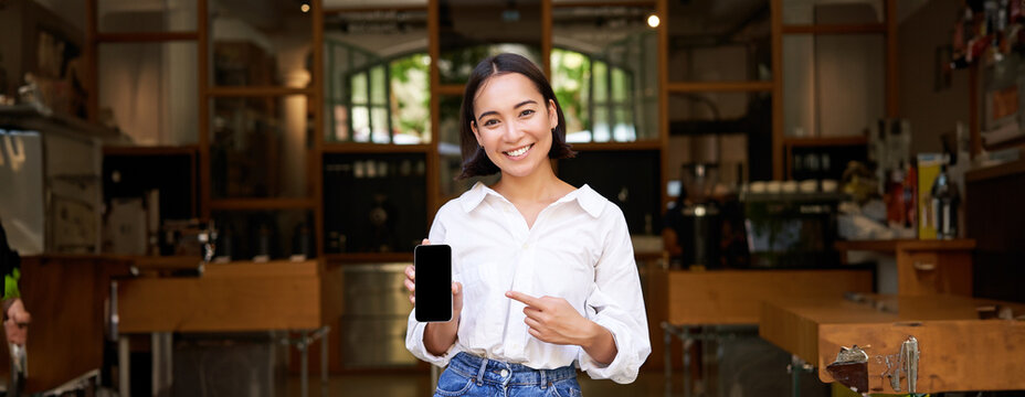 Happy asian woman, cafe manager standing in front of restaurant, pointing finger at smartphone app, showing mobile screen, recommending application