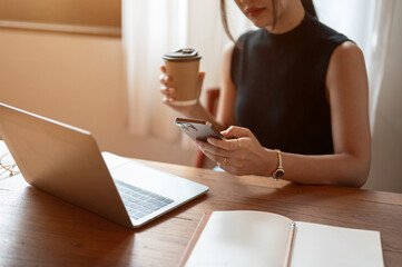 A beautiful Asian woman using her smartphone and sipping coffee while working remotely at a cafe.